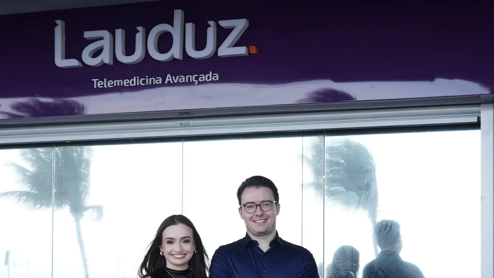 Dr. Wilson Zatt e Dra. Carolina Fernandes Zatt na fachada da nova sede da Lauduz, na orla da Praia de Itaparica, em Vila Velha (ES), sob o letreiro Lauduz — Telemedicina Avançada